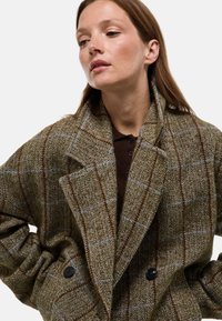 Manteau en laine à chevrons brun et vert avec un devant double boutonnage, de grands revers et des boutons noirs. Présente une coupe décontractée et des épaules structurées.
