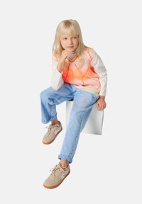 Blond kind dat op een witte blok zit, gekleed in een roze en oranje ruitjesvest, een witte langslavige shirt, lichtblauwe jeans en beige sneakers.
