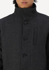 Tmavě šedý kabát se vzorem rybí kosti, vybavený vysokým límcem, zapínáním na knoflíky a boční kapsou. Tkanina má texturovaný vzhled.