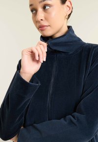 Giacca zip-up in corduroy blu navy con colletto alto, dotata di maniche lunghe e texture a costine. Modella che tiene il colletto con uno sfondo neutro.