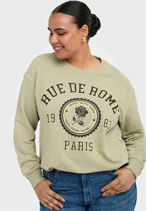 Ung kvinde med mørkt hår samlet i en knold smiler, iført en beige sweatshirt med "RUE DE ROME PARIS 1983" og en rosegrafik samt blå jeans.