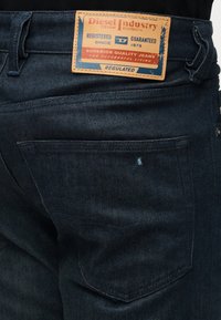 Jeans en denim bleu foncé vue de dos avec une étiquette en cuir marron portant l'inscription "Diesel Industry Division" au-dessus de la poche arrière.
