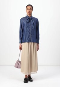 Denim-Hemd mit Knopfverschluss und Fliege, kombiniert mit einem fließenden beigen Rock. Die Frau hält eine kleine lila Handtasche und trägt schwarze Schuhe.