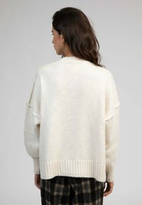 Pull en crème oversized avec un ourlet côtelé. Texture tricot douce, épaules dénudées et légère usure sur les coutures. Pantalon à carreaux visible.