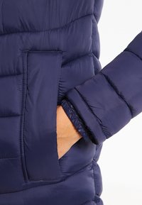 Ärmen på en marinblå vadderad jacka med ribbad mudd och sidoficka, med quiltad struktur och mjukt, lätt material.