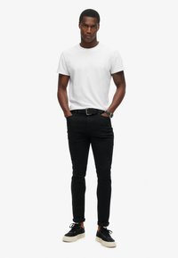 Camiseta blanca de cuello redondo, jeans slim negros y zapatillas negras con suelas blancas. El modelo está de pie con las manos en los bolsillos frente a un fondo simple.