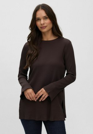 Femme aux longs cheveux bruns portant un haut à manches longues marron foncé et un pantalon foncé, debout devant un fond clair uni.