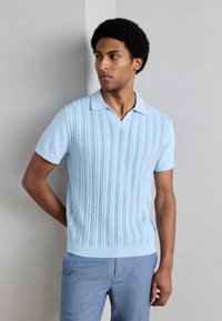 Ανοιχτό γαλάζιο κοντομάνικο πλεκτό πόλο πουκάμισο με V-λαιμόκοψη, διακοσμημένο με υφασμένο κάθετο σχέδιο με καλώδια και ριμπ τελείωμα. Συνδυασμένο με ανοιχτό γαλάζιο παντελόνι.