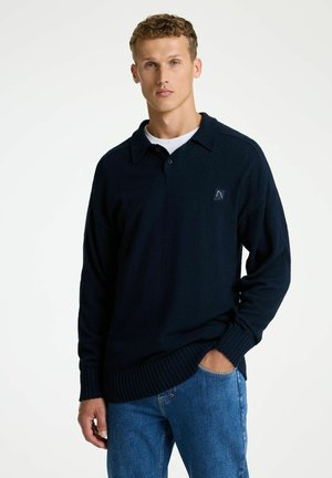 Pull-over bleu marine avec col à revers, manches longues et poignets côtelés. Comprend un petit patch logo sur la poitrine et une coupe décontractée.