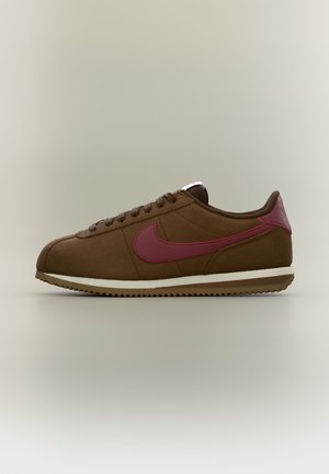 Flacher brauner Sneaker mit burgunderrotem Nike-Swoosh, braunen Schnürsenkeln und gezackter Gummisohle, von der Seite auf neutralem Hintergrund gezeigt.