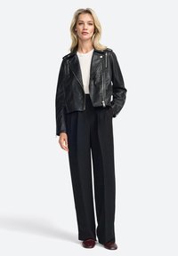 Black leather cropped biker jacket with silver zipper accents, worn over a white shirt and paired with high-waisted black trousers.