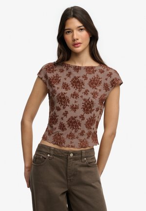 Jeune femme aux longs cheveux bruns portant un haut transparent à manches courtes à motif floral marron et un pantalon taille haute vert olive, debout devant un fond blanc.