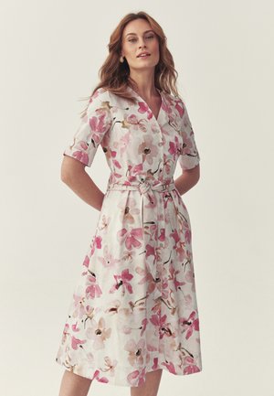 Kobieta w białej sukience do kolan z różowo-beżowym kwiatowym wzorem, z przodu zapinanej na guziki, z wiązanym pasem i rękawami do łokci.