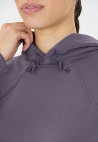 Person som justerar dragsnöret på huvan av en lila hoodie, som visar detaljer av tyget och sömnaden.