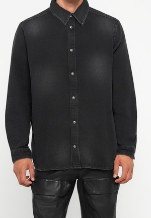 Mężczyzna w wyblakłej czarnej koszuli dżinsowej na guziki z długim rękawem i czarnych spodniach ze skóry, stojący na jednolitym tle.