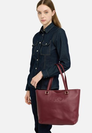 Borsa a tracolla in finta pelle bordeaux con manici doppi, logo impresso e una texture liscia. Il modello indossa una camicia in denim scuro.