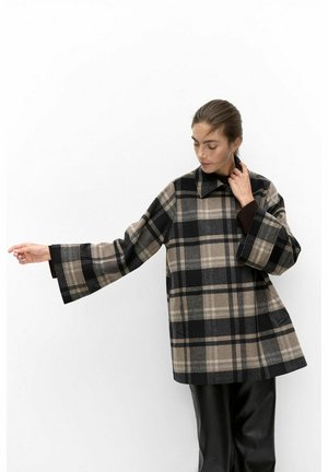 Vrouw die een oversized zwart-beige geruite jas met brede mouwen draagt, staand tegen een effen witte achtergrond.