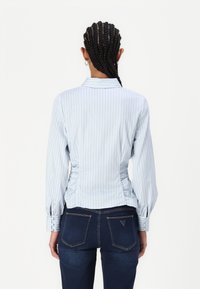 Lyseblå, vertikalt stripete langermet bluse med en tettsittende design, rynkede sider og krage. Parret med mørkeblå jeans.