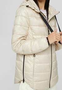 Chaqueta acolchada beige con capucha, con diseño de rombos horizontales, cierre de cremallera negra en los laterales y detalles con cordón.