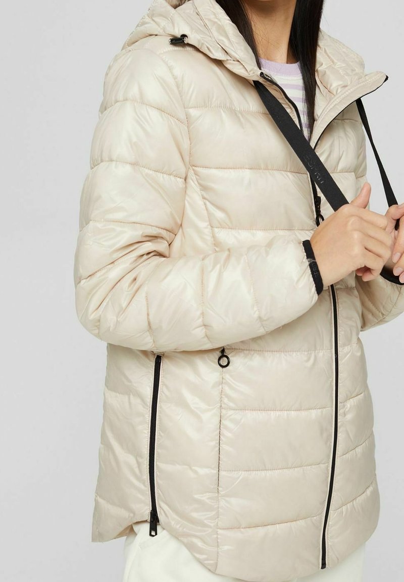 Chaqueta acolchada beige con capucha, con diseño de rombos horizontales, cierre de cremallera negra en los laterales y detalles con cordón.