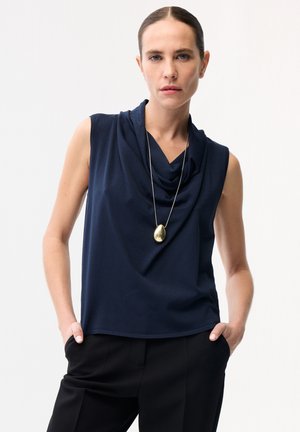 Frau mit zurückgekämmtem Haar trägt ärmelloses, marineblaues Top mit Wasserfallausschnitt, schwarze Hose und lange Halskette mit goldenem Anhänger.