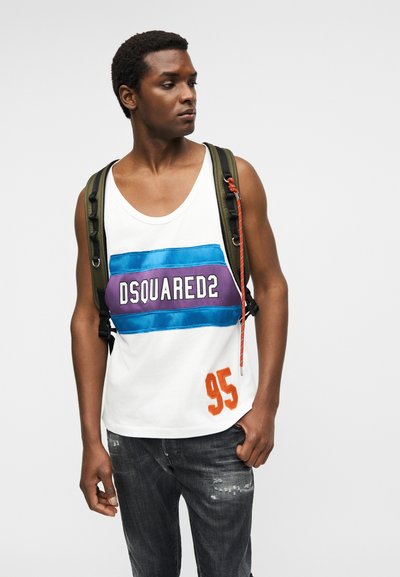 Jeune homme portant un débardeur blanc avec le logo « DSQUARED2 » en bleu et violet, le numéro orange « 95 », un jean foncé et un sac à dos vert, regardant sur le côté.