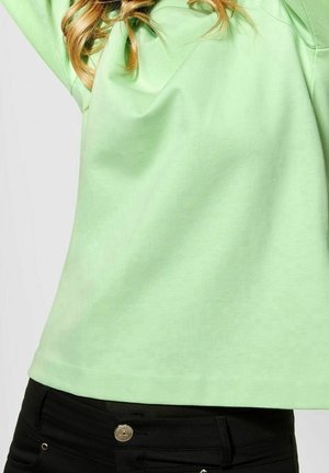 Femme portant un sweat-shirt vert clair et un pantalon noir, levant les bras ; seul le torse et les cheveux sont visibles sur un fond uni.