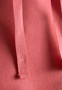 Gros plan sur le tissu d'un sweat à capuche rose avec un cordon de serrage texturé présentant un motif tissé subtil et un petit logo brodé près de l'extrémité.
