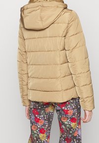 Person iført en beige dunjakke med hætte og farverige bukser med blomsterprint, vist fra ryggen mod en ensfarvet baggrund.