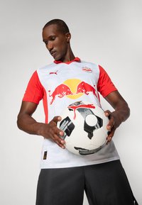 Futbalový dres Puma s bielym trupom a červenými rukávmi, s veľkým žltým kruhom a dvoma červenými býkmi na prednej strane. Čierne šortky a futbalová lopta.