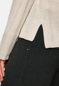 Beige gerippter Pullover mit seitlichem Schlitz, kombiniert mit dunkelgrauen Stoffhosen, die ein rundes metallisches Element an der Tasche aufweisen.