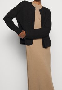 Cardigan nero aperto davanti in maglia testurizzata, indossato sopra un vestito beige. Il cardigan ha maniche lunghe e una vestibilità morbida.