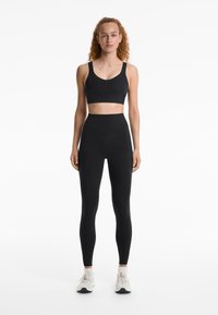 Ensemble de vêtements de sport noir comprenant un soutien-gorge de sport court et un legging taille haute avec une texture lisse. Modèle portant des chaussures de sport blanches.