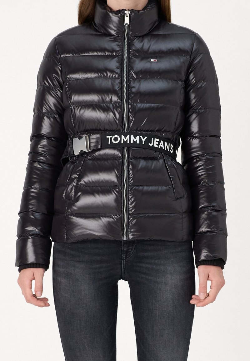 Doudoune noire brillante avec zip devant, poches latérales et une large ceinture noire marquée "TOMMY JEANS", portée avec un jean foncé.