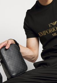 Borsa a tracolla nera in materiale sintetico testurizzato, con un delicato motivo a logo. Può essere portata a mano grazie a una robusta tracolla, dalla forma rettangolare.