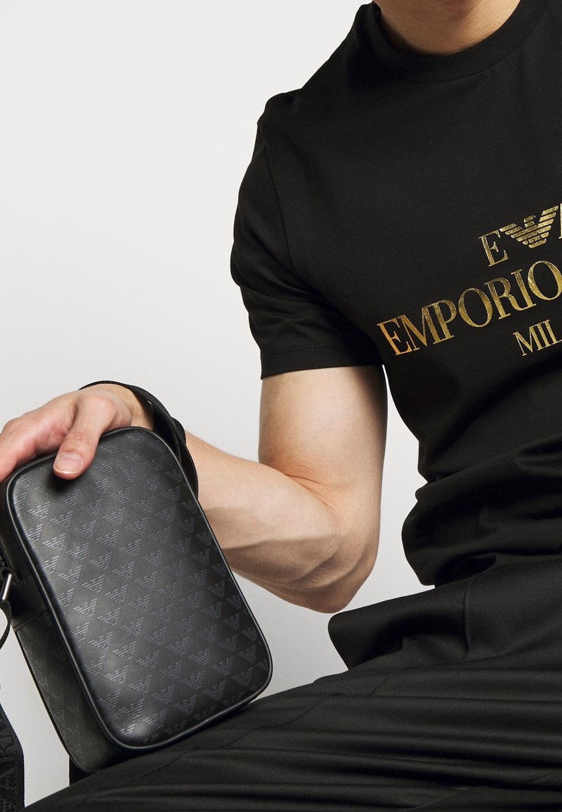 Borsa a tracolla nera in materiale sintetico testurizzato, con un delicato motivo a logo. Può essere portata a mano grazie a una robusta tracolla, dalla forma rettangolare.