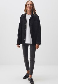 Giacca di jeans nera con bottoni argento, t-shirt bianca, jeans slim scuri e scarpe nere. La giacca ha una vestibilità comoda e due tasche sul petto.