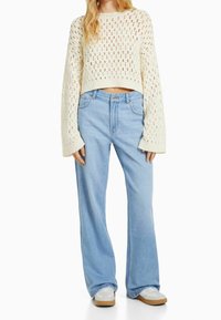 Krämfärgad stickad korttröja med öppet mönster, tillsammans med ljusblå vida jeans. Vita sneakers med bruna sulor fulländar outfiten.