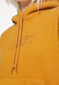 Fleece hoodie i senapsgul med justerbar dragsko. Har en broderad "OBEY"-logga i en subtil kontrastfärg. Mjuk textur.