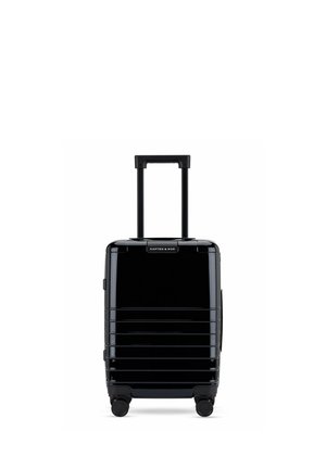 Valise rigide noire avec poignée télescopique, quatre roues pivotantes et rainures horizontales, étiquetée « Kapten & Son » en haut au centre.