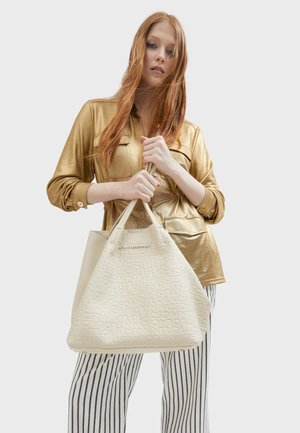 Beige textured tote bag with embossed logo, featuring dual handles. Model wears a shiny gold blouse and striped pants.