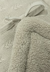 Plaid en laine avec un design texturé en beige clair, un dessous doux et pelucheux, et un ruban en tissu pour la fermeture.