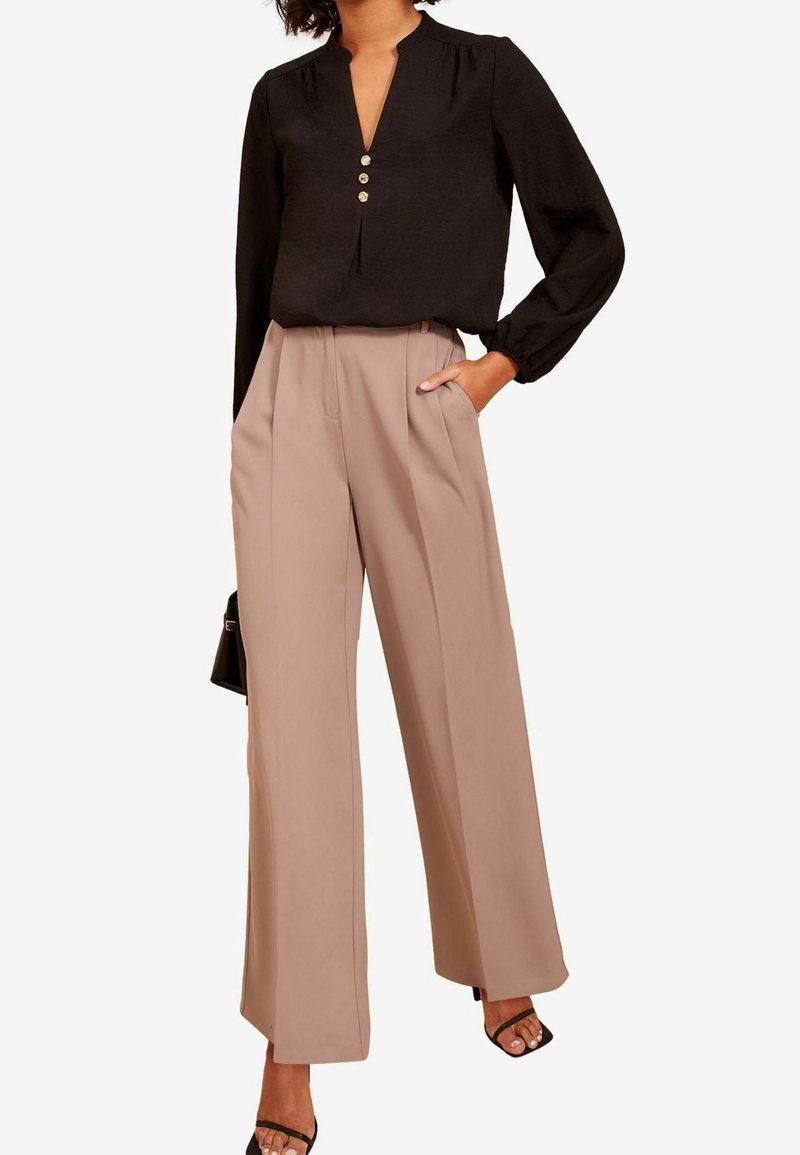 Femme portant un chemisier noir à manches longues avec des boutons et un pantalon beige taille haute à jambes larges, mains dans les poches et sandales noires.
