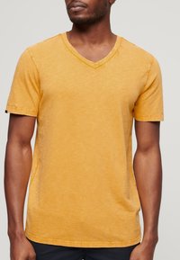 T-shirt a maniche corte, con scollo a V, di un colore giallo senape uniforme, con una finitura texture e dettagli di cucitura discreti.