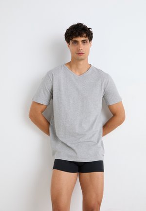 Νεαρός άνδρας με σγουρά σκούρα μαλλιά στέκεται μπροστά σε λευκό τοίχο, φορώντας ανοιχτό γκρι T-shirt με V λαιμόκοψη και μαύρα εφαρμοστά σορτς, με τα χέρια πίσω από την πλάτη.