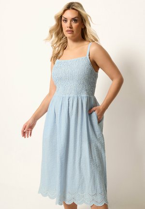 Kvinde med langt blondt hår iført en lyseblå, ærmeløs midi-kjole med smocket bodice og bølget kant, stående med den ene hånd i lommen.