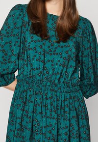 Turkoosi mekko mustalla kukkakuviolla, kerätty vyötärö ja runsaat hihat. Silkkinen kankaan texture, pyöreä pääntie ja elastinen yksityiskohta.