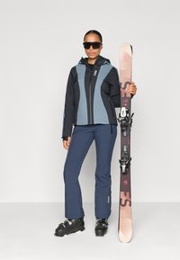 Ski odijelo s crno-plavom jaknom, tamnoplavim hlačama i crnim skijaškim čizmama. Drži skije s ružičastim akcentima i crnim vezovima.