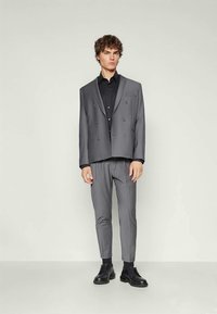 Young man standing in a gray double-breasted suit with black dress shirt and black shoes against a plain light background.