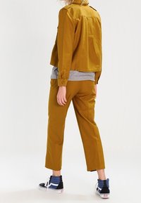 Veste courte jaune moutarde, chemise grise en dessous, pantalon taille haute assorti, et baskets noires avec des accents bleus.